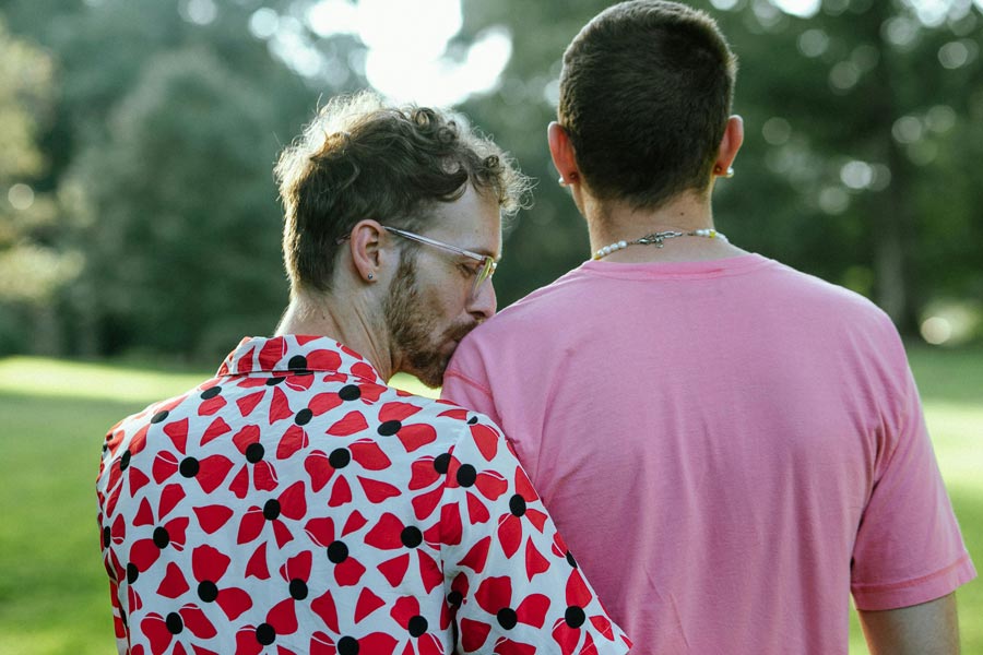 Photo of a gay male couple outside. One of them is kissing the other one's shoulder.