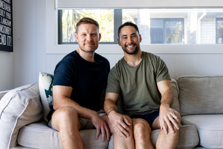 Photo of a gay male couple smiling, sitting on the couch, facing the camera