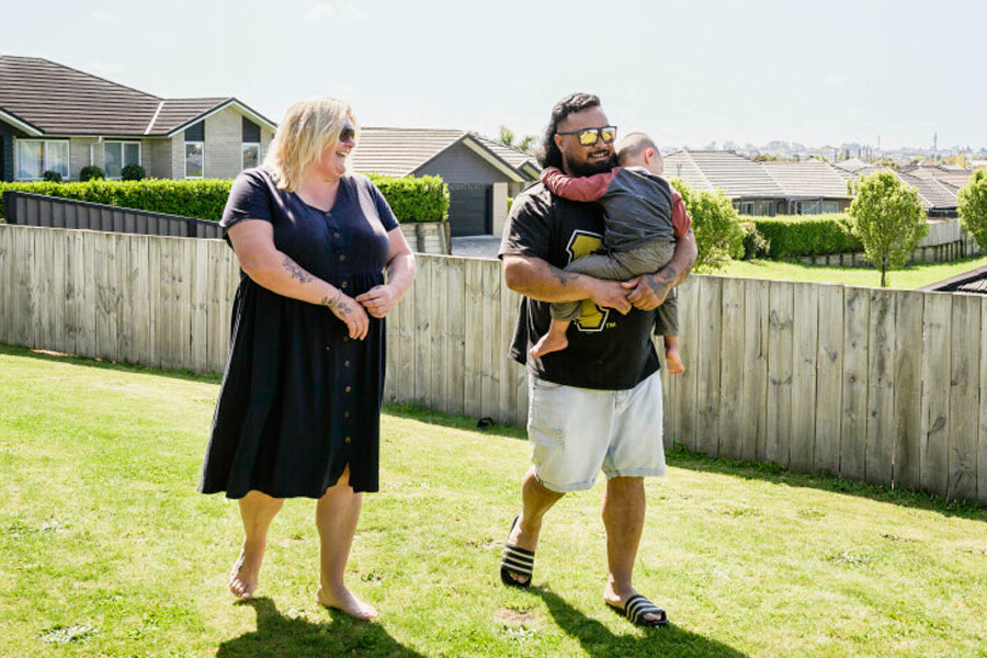 Photo of a happy couple in a Kiwi backyard. The man is holding a baby.